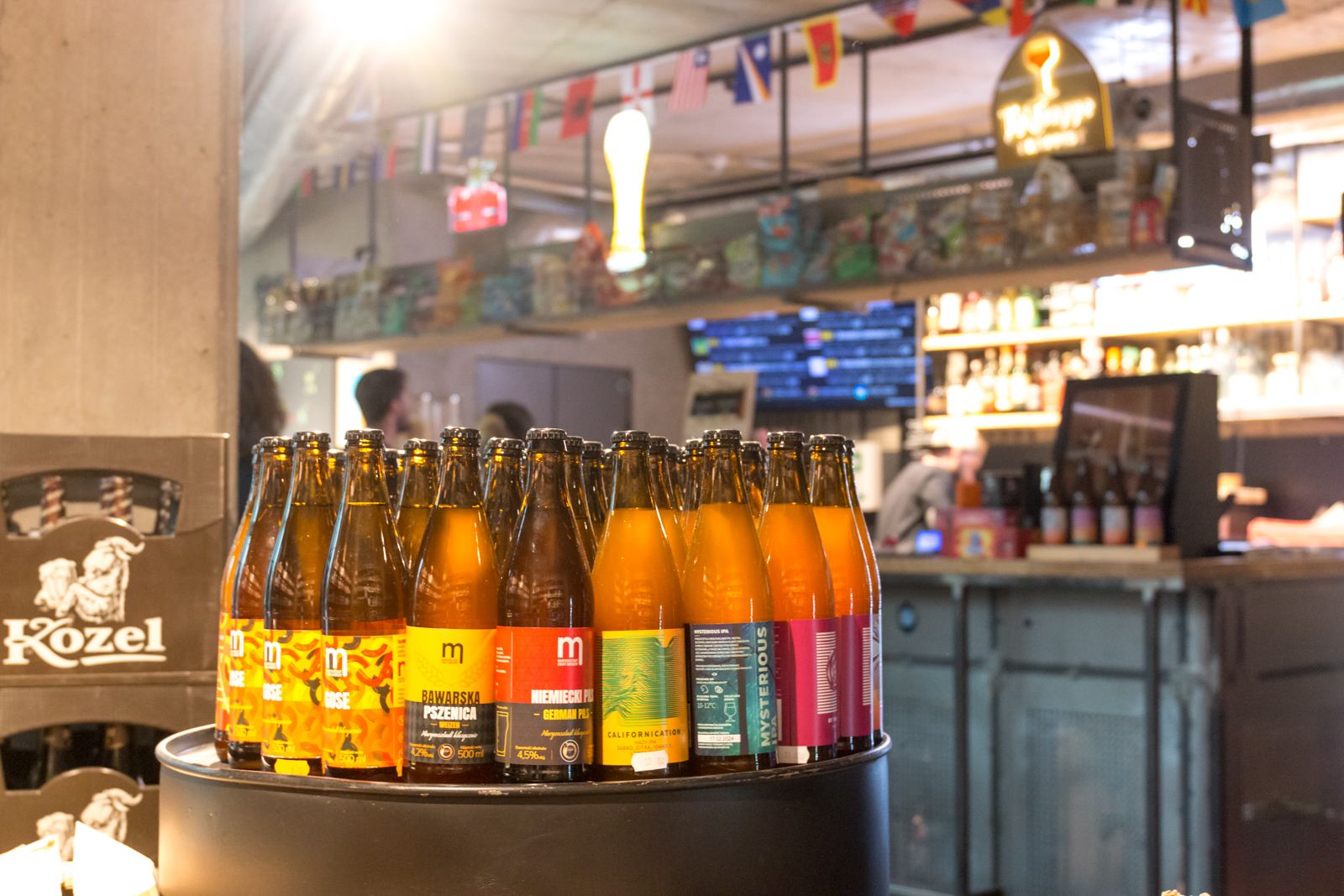 Butelki piwa rzemieślniczego na beczce, w tle bar i ekrany z transmisjami w The Beer Store