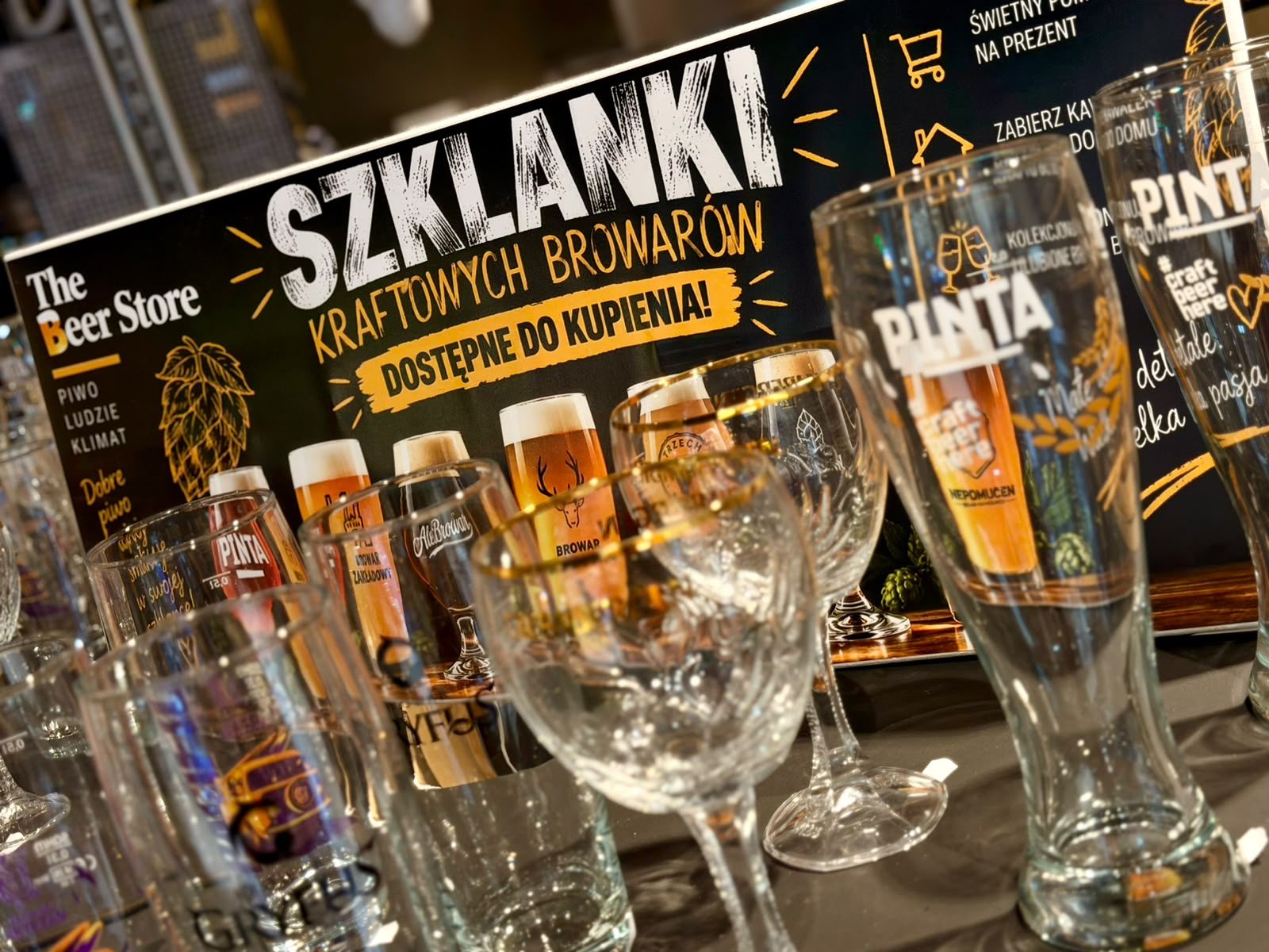 Szklanki, kufle i pokale kraftowych browarów przy plakacie „Szklanki kraftowych browarów dostępne do kupienia” w The Beer Store